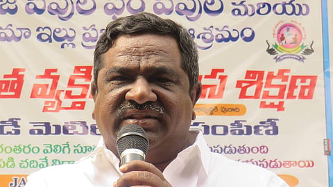 A medium-to-close-up shot of Gade Innaiah, a middle-aged man with dark hair and a mustache, wearing a crisp white button-down shirt. He is holding a microphone with both hands close to his face, appearing to be giving a speech or presentation.

Behind him is a large banner with text in Telugu script. The banner features blue and red lettering on a white background, with a small circular logo on the right side depicting a stylized figure. The man has a serious yet focused expression, and a pen is visible clipped to his shirt pocket. The lighting is bright and even, suggesting an indoor or shaded outdoor event.