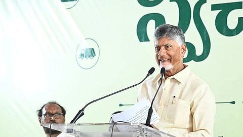 A medium shot shows N. Chandrababu Naidu, the Chief Minister of Andhra Pradesh, standing at a clear acrylic podium and speaking into two microphones. He is wearing a light cream-colored button-down shirt and holding papers in his hand. Behind him is a large white and light green banner with Telugu text and an icon of a garbage truck. Another man in a white shirt and glasses is partially visible sitting to the left in the background.