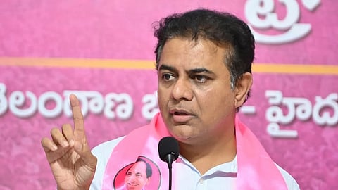 A close-up shot of Indian politician K. T. Rama Rao (KTR) speaking at a podium. He is wearing a light-colored button-down shirt and a bright pink Bharat Rashtra Samithi (BRS) party scarf draped over his shoulders. The scarf features the party's name in multiple languages and a portrait of his father, K. Chandrashekar Rao.

He is captured mid-speech with a serious expression, gesturing with his right index finger raised. In the background, there is a large pink banner with Telugu script. A black microphone stands prominently in the foreground.