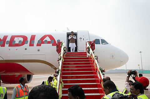 An outdoor, eye-level shot captures a man in white traditional Indian clothing and a brown vest standing at the open door of a white and red Air India plane, with his hands together in a "Namaste" greeting. Red stairs with floral garlands lead down from the plane's door to the tarmac. In the foreground, several people, some in yellow safety vests, are looking toward the plane or taking photos. The background shows a clear sky and the airport's open ground.