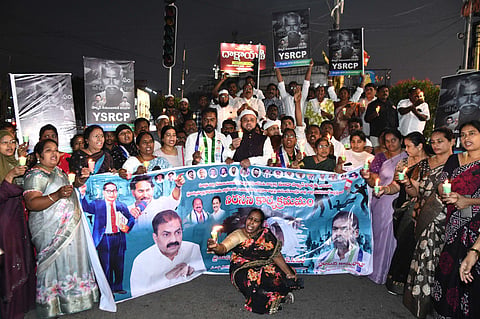 A large group of people, including men and women, are gathered at night for a candlelight vigil and protest. Many participants are holding small, lit candles and posing behind a large, colorful banner.

The banner features several prominent figures, including Y.S. Jagan Mohan Reddy and Dr. B.R. Ambedkar, along with text in Telugu. Individuals in the crowd are also holding up placards with the YSRCP party name and the image of a man. Several men in the center are wearing traditional white clothing and skullcaps, and one man wears a YSRCP party scarf. In the foreground, a woman sits on the ground holding a candle in front of the banner. The background shows a city street at night with a traffic light and a bright billboard.