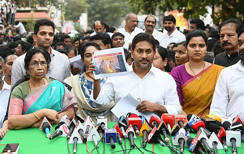 A high-angle, eye-level medium shot shows Y.S. Jagan Mohan Reddy, the former Chief Minister of Andhra Pradesh and leader of the YSR Congress Party, at a press conference. He is standing behind a table covered with a green cloth and crowded with numerous news microphones. He is wearing a white collared shirt and holding a piece of paper in his left hand.

To his right, a woman in a blue and white patterned top holds up a printed photograph for the cameras. To his left, another woman in a purple and orange saree looks toward the camera. An older woman in a green and pink saree is seated to the far left. Several other men and a dense crowd of people are visible in the background, many of whom are looking towards the center of the frame. The lighting is bright and even, suggesting an outdoor daytime setting.