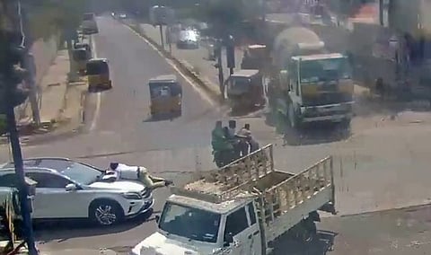 The scene depicts a chaotic moment of traffic:

In the center-left, a white light-duty truck with an open flatbed is angled across the lane.

To the left of the truck, a white SUV is stopped with a person appearing to lean over its hood.

In the center-right, a black motorcycle lies on its side in the middle of the road, suggesting a recent accident.

Various other vehicles are visible, including several yellow auto-rickshaws, a red hatchback, a dark car near the motorcycle, and a large green truck in the background.

The road surface is gray asphalt with faded white lane markings and a zebra crosswalk on the right. Buildings and a few trees line the far side of the intersection.