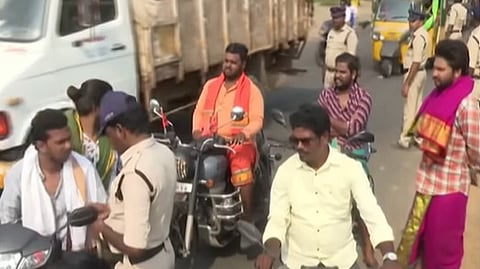 The image shows a busy street scene in Akividu, Andhra Pradesh, likely during a political or religious rally. Several men on motorcycles are being stopped or directed by police officers in khaki uniforms.

In the foreground, a man in a light yellow shirt is riding a motorcycle toward the camera, while behind him, another man wearing an orange shirt and a saffron scarf sits on a Royal Enfield motorcycle. To the right, a man is dressed in traditional attire, wearing a purple and gold dhoti with a matching shawl. A large white truck and a yellow auto-rickshaw are visible in the background, along with other pedestrians and police personnel managing the flow of traffic.