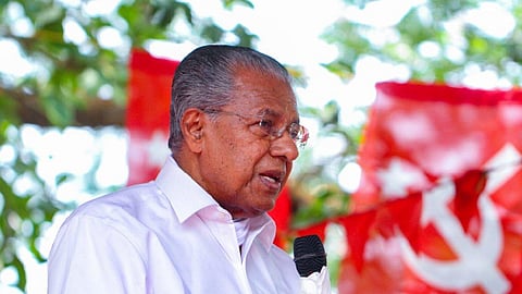 Kerala CPI(M) leader Pinarayi Vijayan speaking into a microphone at a public event, wearing a white shirt and glasses, with red CPI(M) flags blurred in the background.