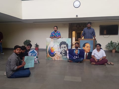 Six students from the Ambedkar Students' Association (ASA) stage a sit-in protest inside a University of Hyderabad administrative building. Centered in the hall, they display a large canvas painting and a framed portrait of B.R. Ambedkar. One student sits holding a purple ASA flag, while another holds a handmade sign reading "HCU Admin Murdabad." The group is positioned in front of office entrances under a wall clock, creating a somber, determined scene of student activism.