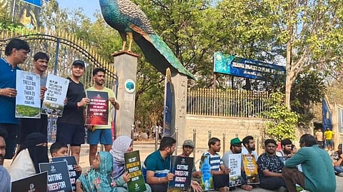 A group of protesters gathers at the entrance of KBR National Park in Hyderabad. Several people sit or stand holding placards with slogans like "Cutting trees for roads?" and "Save KBR." A large peacock statue sits atop the park gate in the background. The image includes "Human Chain" text and hashtags like #SAVEKBR and #SAVETREES, representing a peaceful demonstration by the Parisar Samvad Telangana Chapter against urban deforestation.
