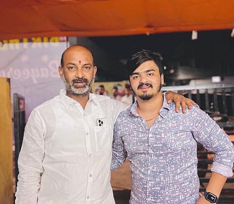 An eye-level medium shot captures two men standing side-by-side. The older man on the left, with a bald head and a graying beard, wears a white button-up shirt and has a red mark on his forehead. He rests his left arm around the shoulder of the younger man on the right, who has dark hair, a mustache, and a goatee. The younger man wears a light blue patterned button-up shirt and a black smartwatch on his left wrist. The background is blurred, showing a warm-toned canopy and some structures.