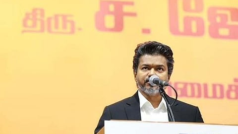 A medium shot of Tamil actor and politician Vijay (right) standing at a wooden podium, speaking into a microphone. He is wearing a black suit over a white shirt and has a focused expression. To his left, an older man with white hair and glasses stands at a matching podium. Both podiums feature a white sign with the letters "PWD" in bold blue and red text and are decorated with vibrant red and pink floral arrangements. The background is a solid, bright yellow-orange wall with partially visible Tamil script in a large pink font. Individuals in formal attire and security are visible in the blurred mid-ground.