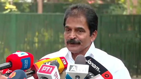 Senior Congress leader KC Venugopal speaking to the media outdoors, surrounded by several microphones from Malayalam news channels during a press interaction. He is wearing a white shirt and appears to be addressing reporters in a calm manner.