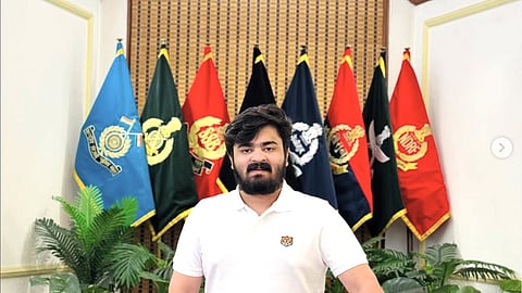 A medium shot of Bandi Bageerath with a dark beard and hair, wearing a white polo shirt with a small tiger emblem, standing in an indoor setting. Behind him is a row of several different colorful flags, including the NDRF (National Disaster Response Force) and SSB (Sashastra Seema Bal). He is positioned between indoor plants and a classic, tufted yellow sofa.