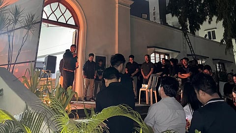 A night memorial service outside a white, Gothic-style church. A speaker stands at the arched entrance while a crowd, mostly in dark attire, listens solemnly. The scene is illuminated by warm interior light, with a "JESUS" sign visible on a nearby building in the background.