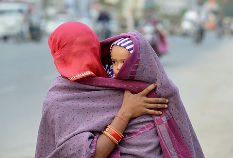 A woman from the back holds a small child who looks over her shoulder. The woman is wearing a red head covering and a purple shawl. The child is wearing a purple-and-white-striped hat and is also wrapped in the shawl. They are standing beside a busy street with people and motor vehicles blurred in the background.