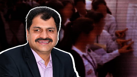 A professional portrait of a man with a mustache, identified as VSN Raju, smiling and wearing a dark blazer over a light purple patterned shirt. He is positioned on the left side of the frame, highlighted with a white outline against a blurred, dark background that appears to show people in an office or classroom setting.