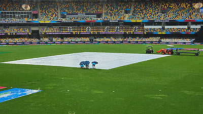 An eye-level, wide shot of an empty cricket stadium in Brisbane, Australia, as seen under a cloudy, overcast sky. At the center of the green field, a white tarp covers the pitch, while a smaller blue tarp is visible in the lower-left corner. Three groundskeepers, huddled under umbrellas, stand on the edge of the large tarp. To the right, a small orange tractor and a utility cart are parked on the grass. The stadium's stands, filled with yellow and blue seats, are largely empty, and the bright stadium lights are on. Banners for the "T20 World Cup Australia 2022" are visible along the perimeter of the field.