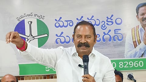 An older South Asian man with dark hair, a prominent thick mustache, and a white collared shirt stands behind a light-colored wooden desk, speaking into a black microphone held in his left hand. His right arm is extended outward to his side with his hand raised in a loose fist gesture. He wears a red string bracelet on his right wrist.

Behind him is a large banner with text written in Telugu script, a green and white logo featuring a ceiling fan, and a partial image of another politician on the right. Two other men are partially visible sitting at the desk below him; one on the bottom left is bald with a thick mustache, looking downwards, and another is partially visible on the bottom right.
