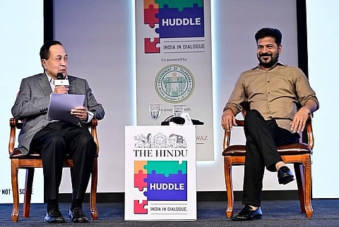 An onstage discussion at "The Hindu Huddle: India in Dialogue" event, co-powered by the Government of Telangana. On the left, N. Ram, Director of The Hindu Publishing Group, wearing a grey blazer and black trousers, holds a microphone and papers while speaking. On the right, A. Revanth Reddy, the Chief Minister of Telangana, sits comfortably in a brown button-up shirt and black trousers, smiling and listening. They are seated in wooden armchairs on either side of a white podium featuring the event's colorful puzzle-themed branding.