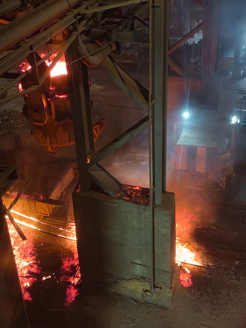 An overhead, high-angle shot of an industrial steel plant accident scene. Molten metal glows intense orange and red as it spills across the dark factory floor. Heavy steel structural beams and a large industrial ladle frame the scene, with smoke and steam rising from the liquid metal.