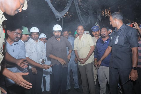 An indoor industrial setting with a hazy, smoky atmosphere. At the center stands Pawan Kalyan, wearing a white safety hard hat and a dark brown shirt, surrounded by a group of men. Several onlookers also wear safety helmets, including a man in a light yellow shirt beside him. Security personnel in uniform and a man in a black shirt stand attentively nearby.