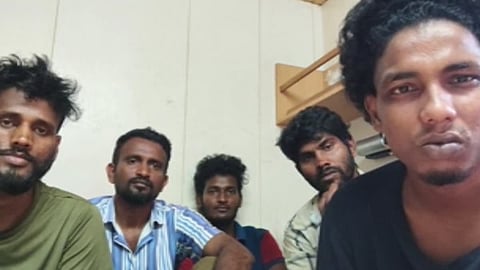 Five distressed young men with dark hair and beards pose closely for a selfie indoors. They look directly at the camera with somber, worried expressions. They are dressed in casual t-shirts and button-downs against a backdrop of plain, off-white wall panels and a simple wooden shelf.