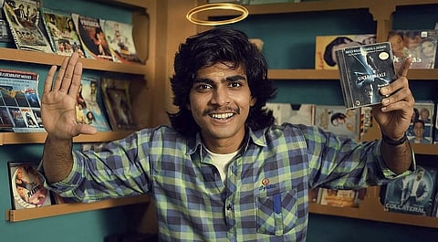 A young man in a checked shirt stands inside a room lined with shelves of CDs/DVDs. He smiles at the camera with one hand raised and holds up a copy of a film/book in the other hand.