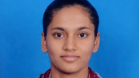 A passport-style studio photograph of a young woman with her hair tied back, posing against a solid blue background. She is wearing a grey, floral-patterned kurta with wooden buttons and a maroon scarf featuring a white geometric design draped around her neck.
