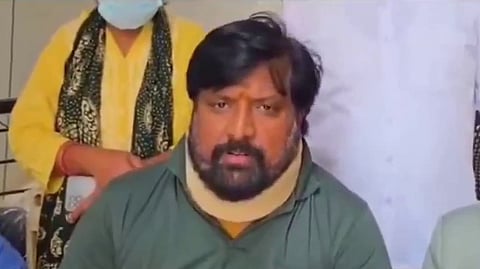 An image showing a close-up shot of a man with dark hair, a prominent mustache, and a thick beard wearing a beige neck brace and a green collared shirt. He appears to be speaking or addressing a group, looking forward with an earnest expression. To his right, another man with short hair and a light green shirt is partially visible, looking down. In the background, two individuals stand behind them—one in a white shirt and another wearing a yellow top with a patterned dark scarf and a light blue medical mask.