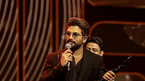 Indian actor Allu Arjun stands on stage at an awards ceremony, holding a microphone in one hand and a Filmfare trophy in the other. He is wearing a black suit, tinted glasses, and a chain necklace, speaking to the audience against a lit, modern backdrop.