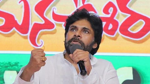 An intense close-up shot of Pawan Kalyan, an Indian actor and politician, speaking passionately into a microphone. He is wearing a white kurta, has a thick beard and mustache, and is raising his right hand in a clenched fist gesture. The background features a vibrant banner with large Telugu script in red against a yellow and green backdrop.
