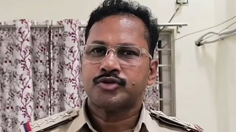An Indian police officer, seen from the chest up, is wearing a khaki uniform with three stars on his shoulder epaulets. He is a middle-aged South Asian man with short black hair, glasses, and a prominent mustache, appearing to speak into an attached lapel microphone. The background consists of a plain light-colored wall and a patterned floral curtain.