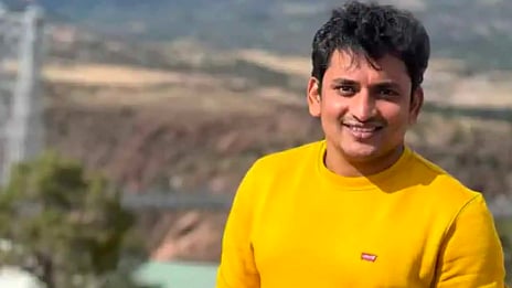 A smiling man wearing a bright yellow sweatshirt stands outdoors with hills and a blurred landscape in the background.