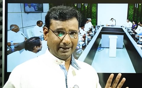 An Indian journalist or commentator wearing thin-rimmed glasses and a white button-up shirt with a blue inner collar standing in front of a large monitor screen. The screen behind him displays a video or image of a formal meeting or conference room setting, where several officials are seated around a large U-shaped wooden conference table.