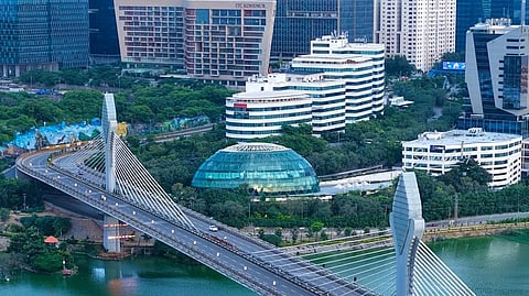 An aerial, vertical shot shows the Durgam Cheruvu Cable Bridge spanning across a calm, green lake in Hyderabad. The bridge features two large, sleek grey concrete pylons from which numerous white stay cables extend to support the multi-lane roadway. A few vehicles are visible on the bridge.

In the background, a dense urban skyline rises behind lush green trees. Notable buildings include a distinctive glass dome structure nestled in the greenery, the curved facade of the ITC Kohenur hotel, and several modern, high-rise glass corporate towers under an overcast sky.