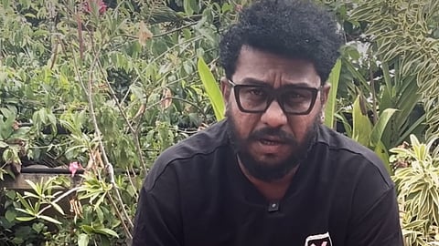 A close-up shot of a man with thick curly hair, a beard, and prominent black-rimmed glasses, speaking directly toward the camera. He is wearing a plain black t-shirt with a small graphic logo on the lower left side. The background consists of dense, lush green plants and foliage.