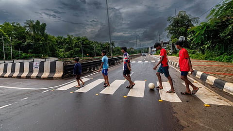 Five children walking a zebra crossing on a rain-soaked road. One among them nudging a football forward. 