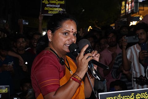 Kavitha, wearing an orange saree and maroon blouse, speaks into a microphone at a nighttime public gathering as supporters stand behind her holding placards and recording on their phones.