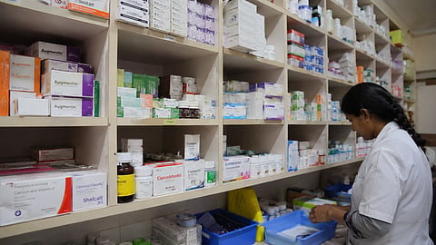 A pharmacist in a white coat sorts medicines in a pharmacy, with shelves stocked with medicine boxes and bottles in the background.
