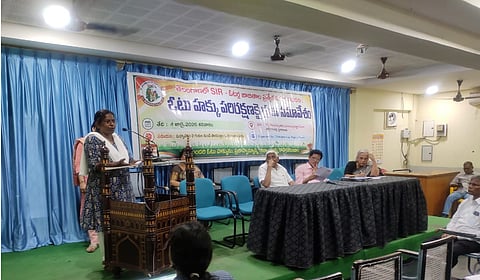 A speaker addresses a public meeting organised by the Telangana Voter Rights Forum at a hall in Hyderabad, while panellists sit on stage beneath a banner about the Election Commission's Special Intensive Revision (SIR) exercise. Attendees are seated in front of the stage.
