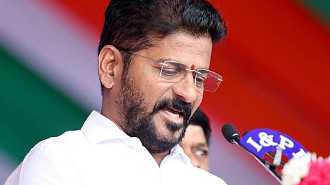 A medium close-up shot of an Indian man with short black hair, a beard, and a mustache, wearing rimless eyeglasses and a crisp white collared shirt. He is leaning slightly forward at a dark wooden podium, looking downward as if reading a speech or document. A microphone with an "I & PR" logo is positioned near him, and a small pen holder sits on the edge of the podium. The background features a blurred red, white, and green banner, with a small portion of another person's face visible behind him.