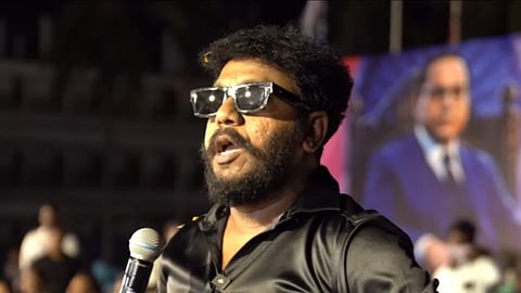 An Indian man with a full beard, mustache, and short, curly black hair stands outdoors at night speaking into a microphone. He wears clear-rimmed black sunglasses and a black button-down satin shirt. In his right hand, he holds a silver and black microphone up toward his mouth, which is open as if speaking or shouting. The background is blurred, showing an outdoor audience at night and a large screen or banner featuring a portrait of B. R. Ambedkar.
