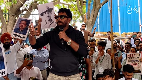 Raavan speaks into a microphone at an outdoor protest. He wears a black hoodie and sunglasses, gesturing with his hand. Surrounding protestors hold signs featuring historical and political figures, including Bhagat Singh, alongside a placard reading "NEET PAPER LEAK."