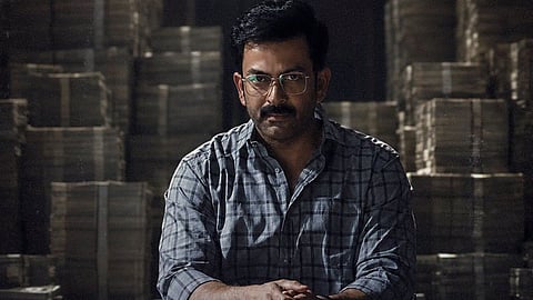 Prithviraj Sukumaran as Rajeevan in a scene from I, Nobody, seated with his hands clasped, against a dimly lit backdrop stacked with bundles of cash.