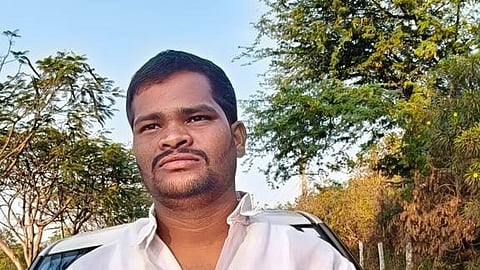 A medium close-up shot of a man standing outdoors in front of a white vehicle. The man has short, dark hair, a mustache, and a light beard. He is wearing a white, short-sleeved, button-up shirt and is looking slightly upward and to the right with a neutral expression. The background features green trees, foliage, and a clear blue sky, suggesting an outdoor setting during the daytime.