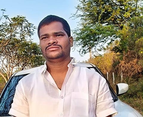 A medium close-up shot of a man standing outdoors in front of a white vehicle. The man has short, dark hair, a mustache, and a light beard. He is wearing a white, short-sleeved, button-up shirt and is looking slightly upward and to the right with a neutral expression. The background features green trees, foliage, and a clear blue sky, suggesting an outdoor setting during the daytime.
