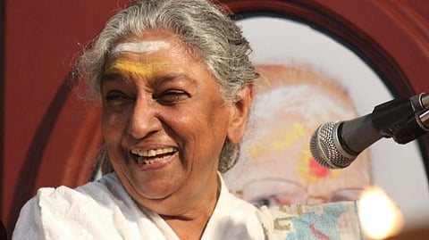 Veteran playback singer S Janaki smiles while seated beside a microphone during a public event, wearing a white sari and looking away from the camera.