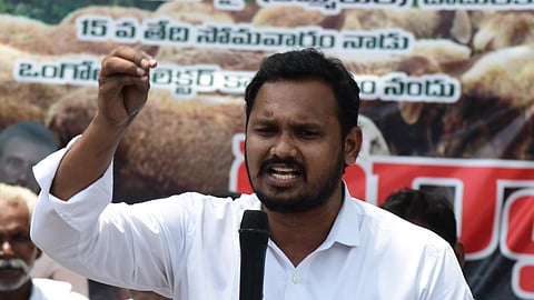 An intensive medium shot captures a man with a beard and mustache wearing a white collared shirt, holding a microphone in his left hand and gesturing with his right. He speaks passionately in front of a banner featuring Telugu text, with a crowd seated behind him.