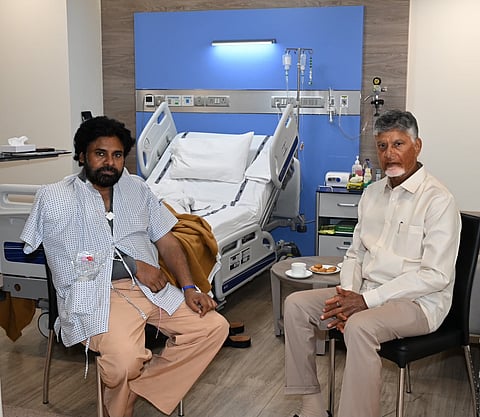 A photograph taken inside a modern hospital room shows two prominent Indian politicians, Pawan Kalyan and N. Chandrababu Naidu, sitting next to each other.

On the left, Pawan Kalyan is seated on a chair, wearing a patterned hospital gown over light tan trousers. He has a full beard and an IV line or medical device secured to his arm. On the right, N. Chandrababu Naidu sits on a black office chair, dressed in his characteristic light beige button-up shirt and matching trousers.

Behind them is an empty, adjustable hospital bed with white sheets and a brown blanket slouched on it, set against a blue accent wall equipped with medical outlets, an IV pole, and a light fixture. A small round table sits between them holding a cup and saucer alongside a plate of biscuits.