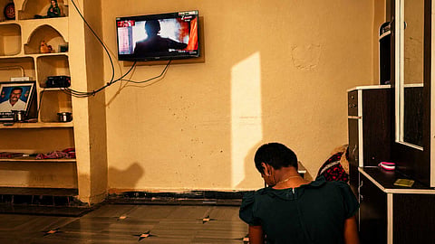 A person with short hair, wearing a dark green top, sits on the floor with their back to the camera inside a room. In front of them, a flat-screen television mounted on a plain wall displays a Telugu news or entertainment channel. To the left is a built-in shelving unit with household items, and to the right is a wooden wardrobe with a mirror.
