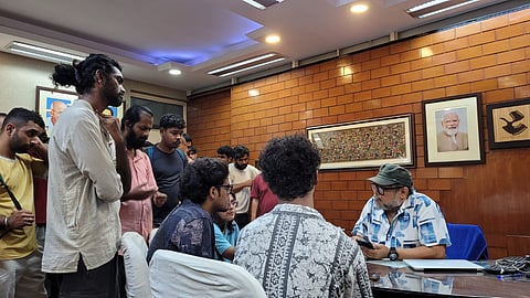 Several SRFTI students stand and sit around a table as they meet Vice Chancellor Samiran Datta in his office to discuss the delay in disciplinary action following an Internal Committee's sexual harassment findings. Framed photographs and artwork are visible on the office wall.