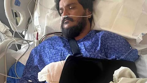 An close-up shot shows an adult male of South Asian descent lying in a hospital bed with his eyes closed. He has a prominent dark beard and mustache. He is wearing a blue hospital gown with a repeating pattern and is hooked up to medical equipment, including a nasal cannula providing oxygen. His left arm is wrapped in thick white bandages and supported by a black sling across his chest, while an IV line is visible in his right hand.
